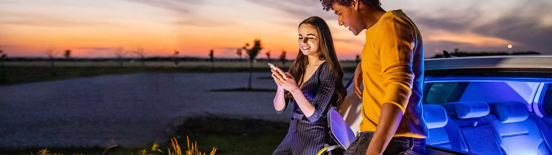 Zwei junge Erwachsene stehen bei Sonnenuntergang an einem Auto und schauen gemeinsam auf ein Smartphone, das Aufladen eines Elektroautos ist im Hintergrund sichtbar.