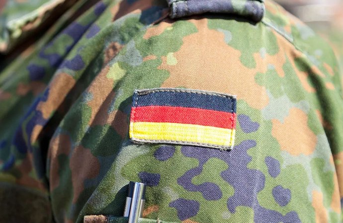 Nahaufnahme eines Bundeswehrsoldaten in Flecktarnuniform, auf dessen Ärmel ein Aufnäher mit der deutschen Flagge befestigt ist. Darunter steckt ein Stift in einer kleinen Tasche, die Sonne wirft Licht auf den Stoff.