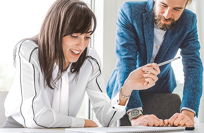Junge Frau sitzt l&auml;chelnd an einem Schreibtisch und zeigt auf ein Dokument. Ein Mann steht neben ihr und blickt ebenfalls auf das Papier. Die Szene wirkt freundlich und beratend.