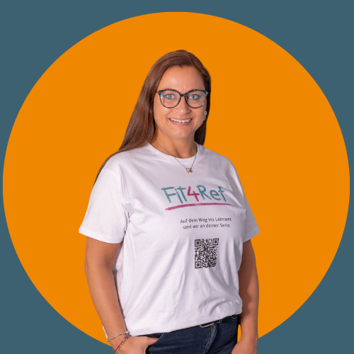 Frau mit Brille und langen braunen Haaren steht vor orangem Hintergrund. Sie tr&auml;gt ein wei&szlig;es T-Shirt mit &bdquo;Fit4Ref&ldquo;-Logo und QR-Code und l&auml;chelt in die Kamera.