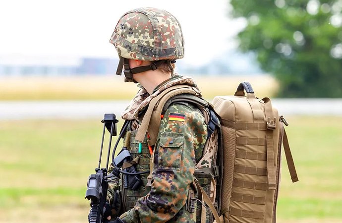 Soldat der Bundeswehr in Tarnuniform mit Helm und großem Rucksack, seitlich stehend mit Gewehr auf freiem Gelände.