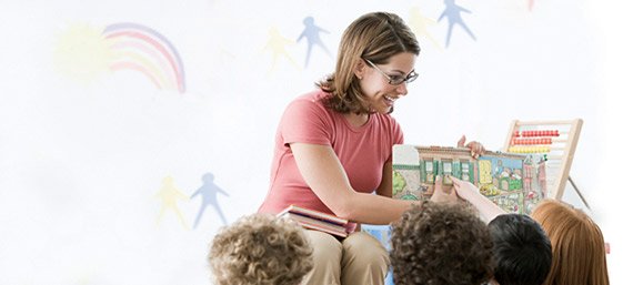 Wichtige Versicherungen für Lehrer der DBV Deutsche Beamtenversicherung der Absolut Finanz in Ulm Eine Frau mit Brille und rosa T-Shirt sitzt vor einer Gruppe kleiner Kinder und zeigt ihnen ein Bilderbuch. Die Kinder schauen aufmerksam auf die Buchseiten. Im Hintergrund sind bunte Wandmalereien mit Regenbogen und Figuren sowie ein Holzstuhl mit Abakus zu sehen.