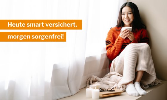 Eine Frau mit schwarzen Haaren, einem roten Pulli und einer Decke über den Knien, sitzt in der Ecke auf einer Fensterbank und trinkt einen Tee aus einer weißen Tasse.