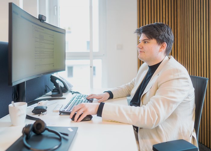 Büroarbeit am Computer Ein Mann im hellen Sakko sitzt an einem Schreibtisch im Büro und arbeitet konzentriert an einem Computer mit großem Bildschirm. Neben ihm stehen eine Kaffeetasse, ein Telefon und ein Headset.