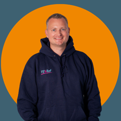 Hannes Hoppe steht vor orangem Hintergrund und l&auml;chelt. Er tr&auml;gt einen dunkelblauen Hoodie mit Fit4Ref-Logo und hat die H&auml;nde locker in den Taschen.