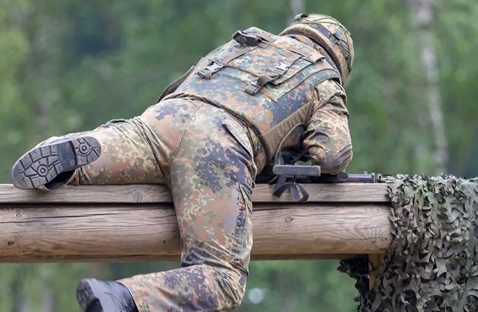 Ein Soldat in kompletter Tarnuniform und Schutzweste liegt bäuchlings auf einem Holzsteg. Er hält sein Gewehr im Anschlag und zielt konzentriert, während neben ihm ein Tarnnetz hängt und im Hintergrund unscharf Bäume zu sehen sind.