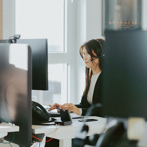 Mitarbeiterin im Büro am Computer Eine Frau mit Headset sitzt im Büro an einem Schreibtisch und arbeitet an zwei großen Monitoren. Vor ihr liegen Telefon und Unterlagen.