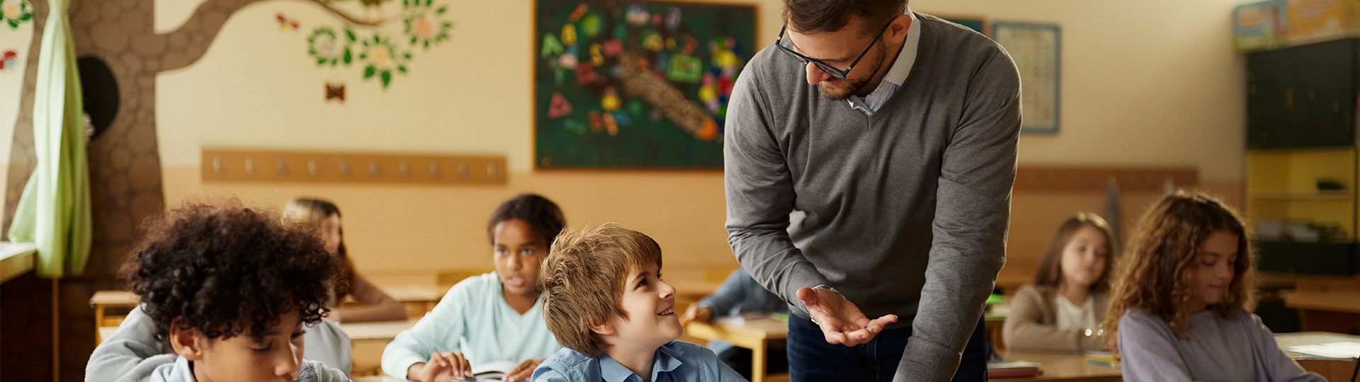 Ein Lehrer beugt sich &uuml;ber einen Sch&uuml;lertisch und spricht mit einem l&auml;chelnden Jungen. Im Hintergrund sitzen weitere Kinder in einem bunt dekorierten Klassenzimmer.