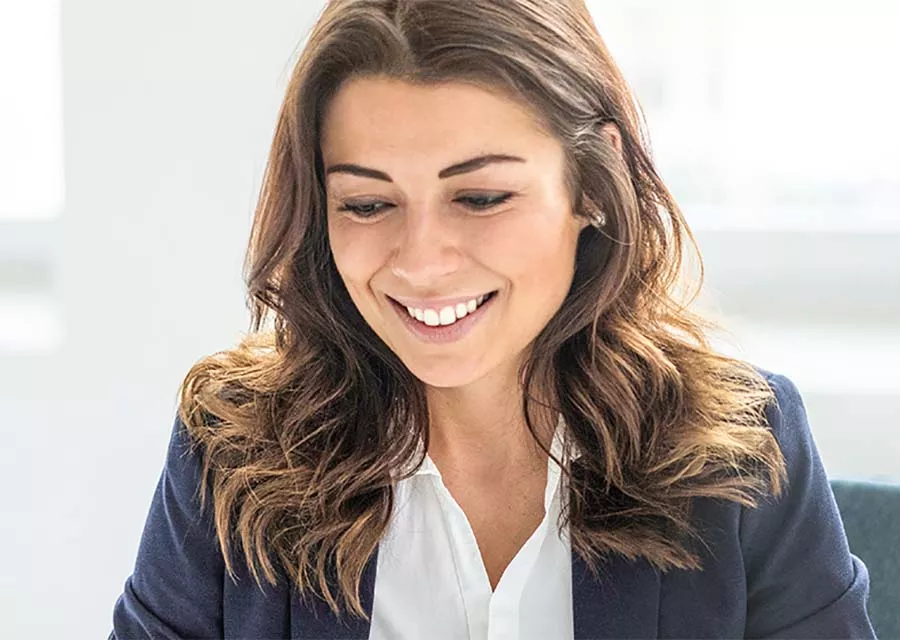 Junge Frau mit braunen, welligen Haaren und Business-Outfit sitzt lächelnd am Schreibtisch in einem hellen Büro. Sie blickt nach unten, wirkt zufrieden und konzentriert.