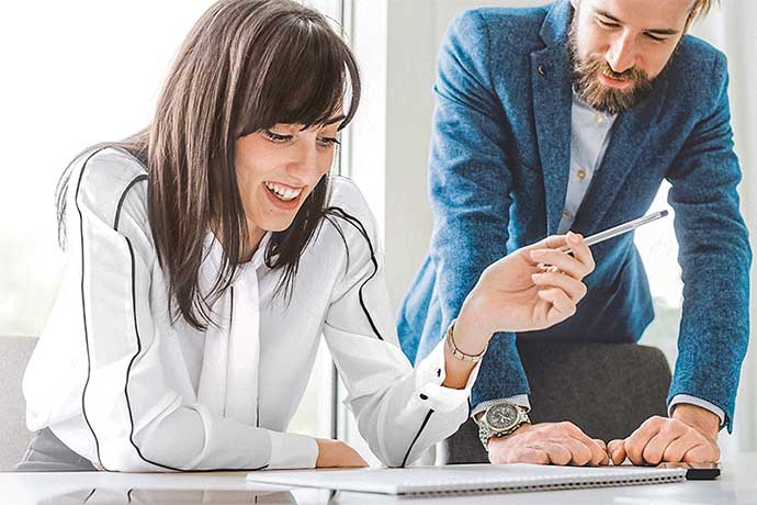Junge Frau sitzt l&auml;chelnd an einem Schreibtisch und zeigt auf ein Dokument. Ein Mann steht neben ihr und blickt ebenfalls auf das Papier. Die Szene wirkt freundlich und beratend.