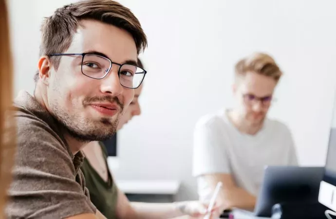 Wichtige Versicherungen für Verwaltungsbeamte der DBV Deutsche Beamtenversicherung der Absolut Finanz in Ulm Ein junger Mann mit Brille blickt lächelnd in die Kamera, während zwei weitere Personen konzentriert an Laptops arbeiten. Die Szene spielt in einem modernen, hellen Büro.