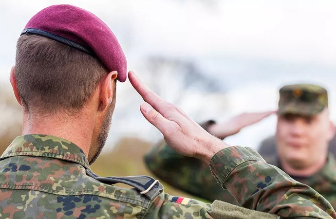 Ein Bundeswehrsoldat in Flecktarnuniform und bordeauxrotem Barett salutiert. Im Hintergrund stehen zwei weitere Soldaten, von denen einer ebenfalls salutiert.