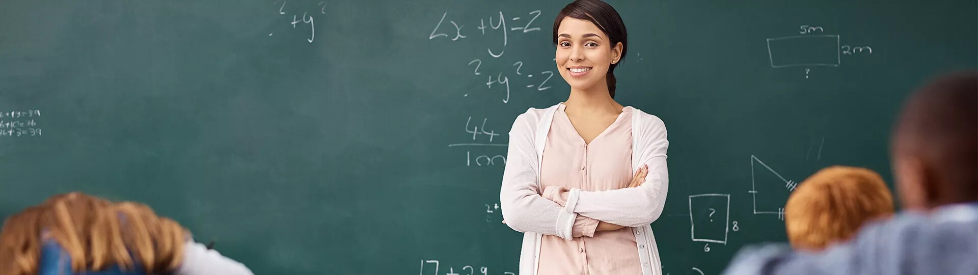 Eine Lehrerin steht mit verschränkten Armen vor einer Tafel mit mathematischen Formeln. Im Vordergrund sitzen mehrere Schülerinnen und Schüler.