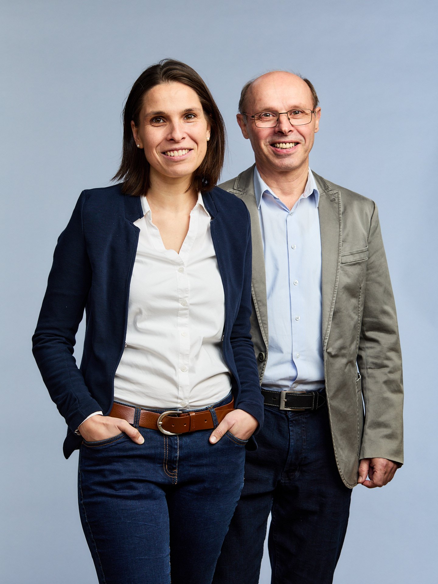 Zwei l&auml;chelnde Personen stehen nebeneinander vor hellem Hintergrund, eine Frau mit dunklem Blazer und wei&szlig;er Bluse, die H&auml;nde in den Hosentaschen, und ein Mann mit Brille, Hemd und Sakko.