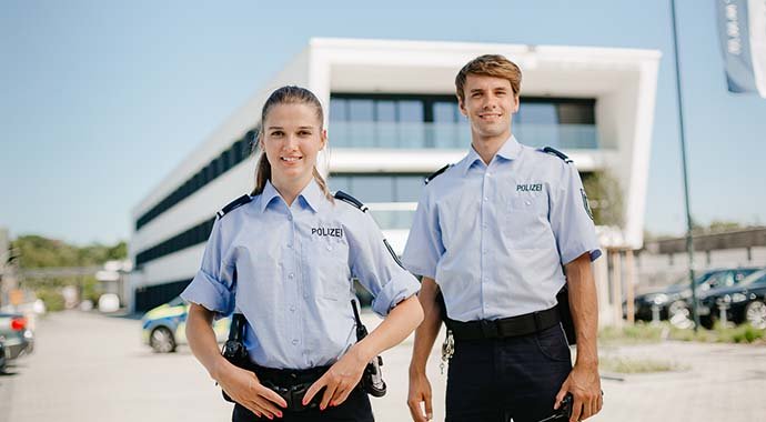 Eine Polizistin und ein Polizist in hellblauer Uniform stehen l&auml;chelnd vor einem modernen B&uuml;rogeb&auml;ude. Beide tragen G&uuml;rtel mit Ausr&uuml;stung und wirken freundlich und professionell.