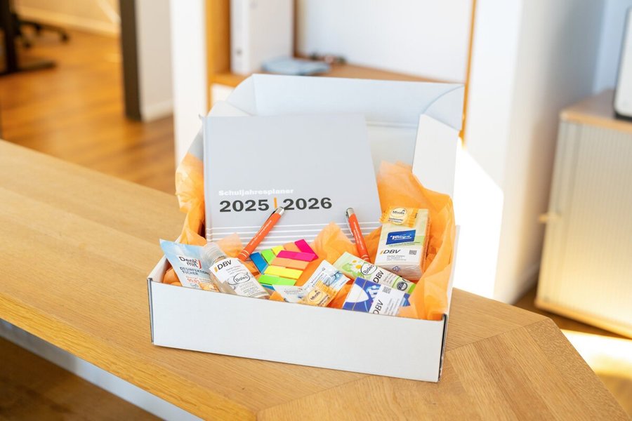 DBV-Starterpaket der DBV Deutsche Beamtenversicherung Stefan Greten in Cloppenburg Eine geöffnete Geschenkbox steht auf einem Holztresen. In der Box liegen verschiedene Büroartikel wie Textmarker, Stifte und Haftnotizen, umgeben von orangefarbenem Papier. Auf einem Heft in der Box steht die Jahreszahl „2025–2026“.