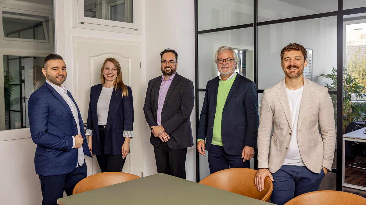 Fünf Mitarbeiterinnen und Mitarbeiter eines Versicherungsteams stehen nebeneinander in einem modernen Büro mit Glastrennwänden. Alle tragen Businesskleidung und lächeln in die Kamera. Vor ihnen steht ein Besprechungstisch mit Stühlen.