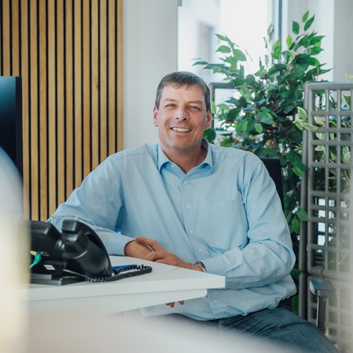 Krankenversicherung für Beamte Ein Mann im blauen Hemd sitzt entspannt an einem Schreibtisch im Büro und lächelt in die Kamera. Neben ihm steht ein Telefon, im Hintergrund sind Pflanzen zu sehen.